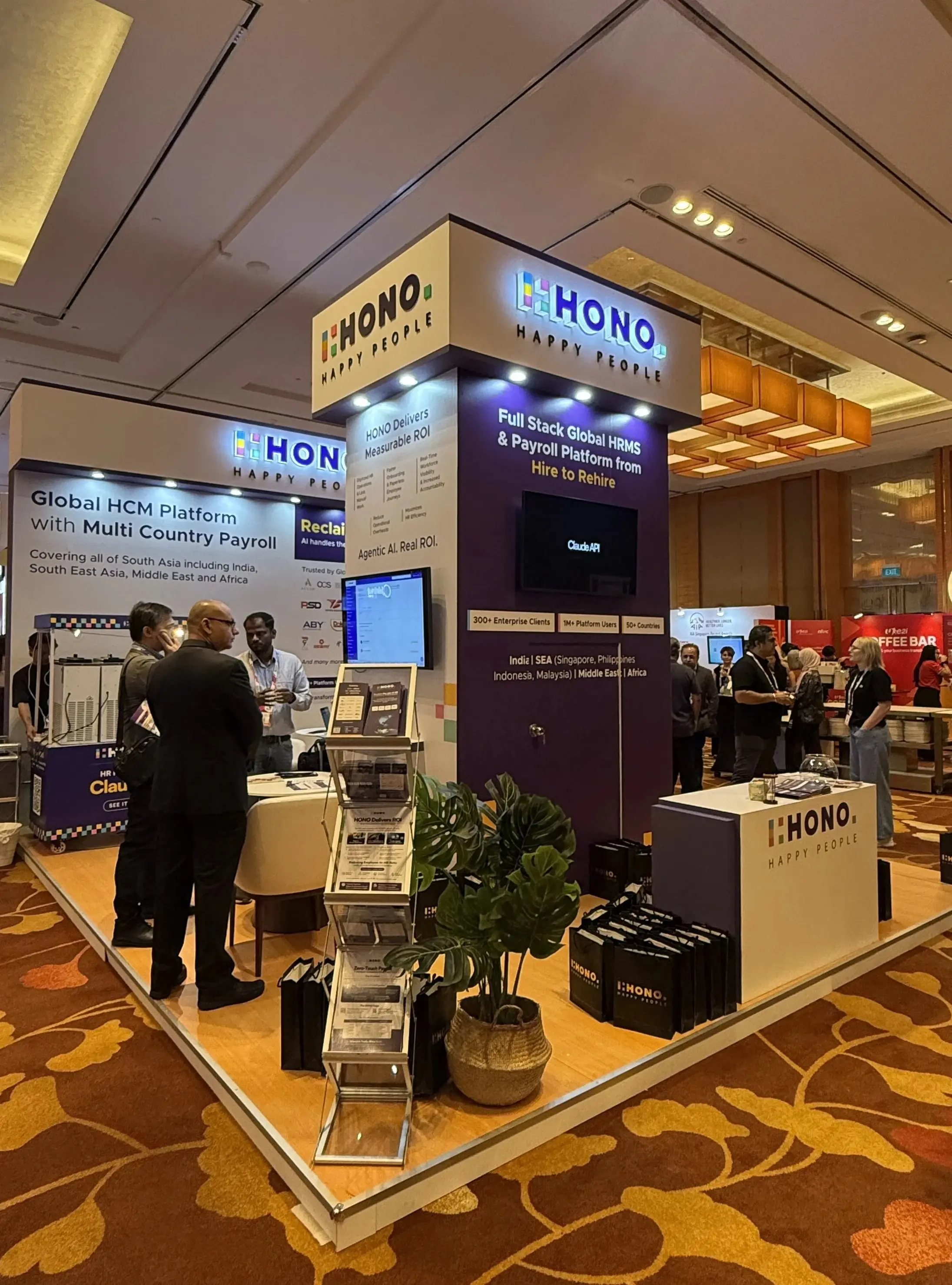 HONO booth at a PeoplesMatter Tech HR Singapore featuring HRMS and payroll solutions, with branded displays, demo screens, brochures, and attendees engaging in conversation around the stand.